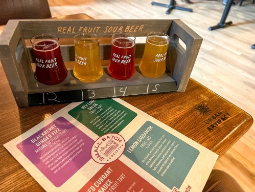 A wooden flight holder with four colorful sour beer samples and a menu on a wooden table.