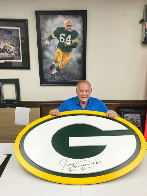 A man sits with a large signed Green Bay Packers logo, smiling in front of a framed photo of a player.