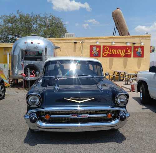 A classic black 1957 Chevrolet parked in front of a food trailer labeled "Jimmy's" under a clear blue sky.