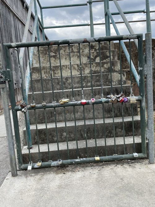 A metal gate with multiple colorful padlocks attached, leading to a set of stairs. The background is cloudy.