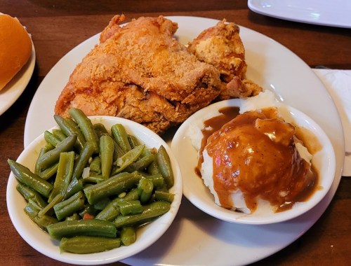 A plate with fried chicken, green beans, and mashed potatoes topped with gravy. A roll is visible in the background.