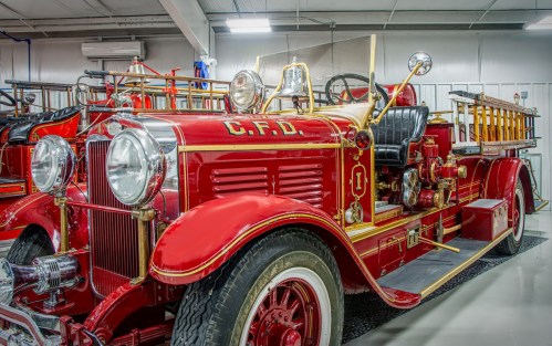 A vintage red fire truck with gold detailing, featuring a bell and ladder, displayed in a garage.