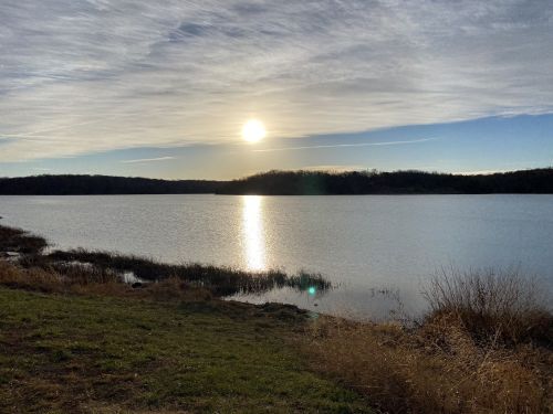 A serene lake at sunset, with the sun reflecting on the water and a grassy shoreline in the foreground.