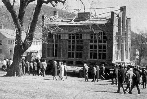 Historic photo of a building being moved on a platform, with a crowd watching and trees in the background.