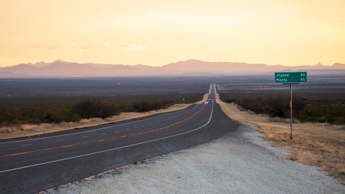 A long, straight road stretches into the distance, with mountains and a sunset in the background. Signpost indicates distances.