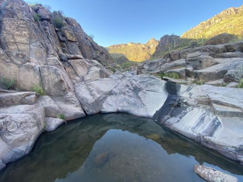 A serene rock formation surrounds a calm pool of water, with mountains in the background under a clear blue sky.