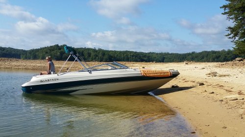 A person sits on a boat beached on a sandy shore, surrounded by trees and a clear blue sky.