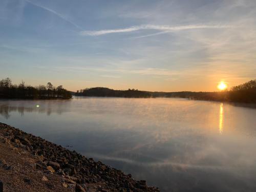 A serene lake at sunrise, with mist rising from the water and a rocky shoreline under a clear blue sky.