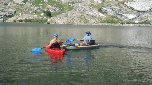 Two people in kayaks on a calm lake, one in a red kayak and the other in a small boat, surrounded by rocky hills.