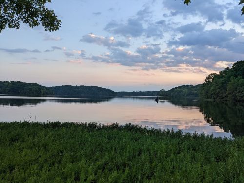 A serene lake at sunset, reflecting clouds and trees, with a lone figure in a boat on the water.