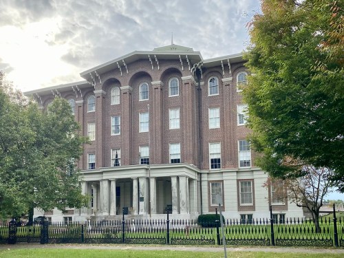 Historic brick building with large columns, surrounded by trees and a black fence, under a cloudy sky.