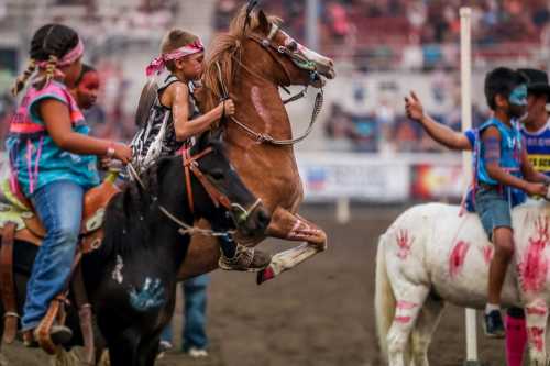 Children participate in a rodeo, riding horses with colorful face paint and vibrant attire in a lively arena.