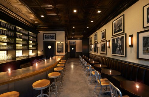 A stylish bar interior with a long counter, wooden stools, and framed black-and-white photos on the walls.