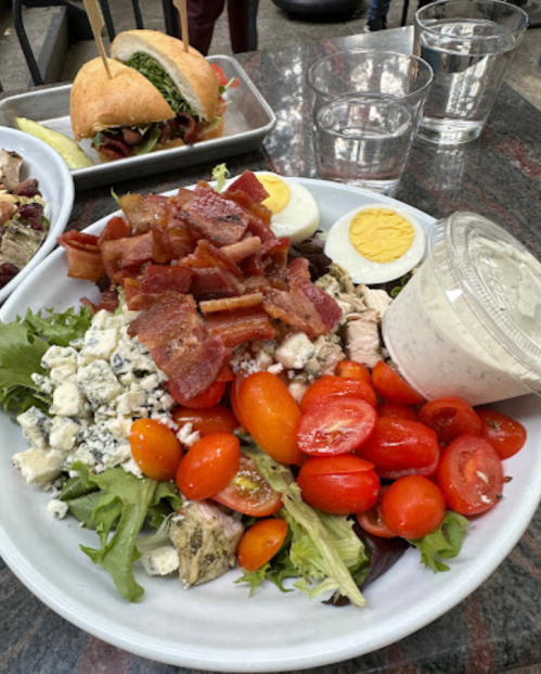 A large salad with greens, cherry tomatoes, bacon, hard-boiled eggs, chicken, blue cheese, and a dressing on the side.