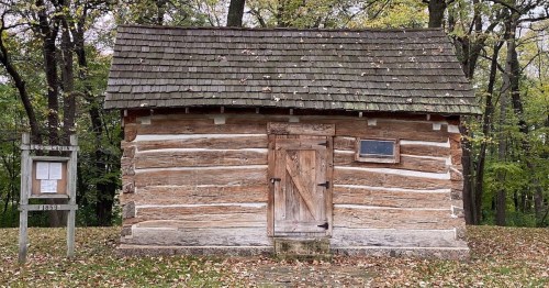 A rustic log cabin with a wooden door and a shingled roof, surrounded by trees in a wooded area.