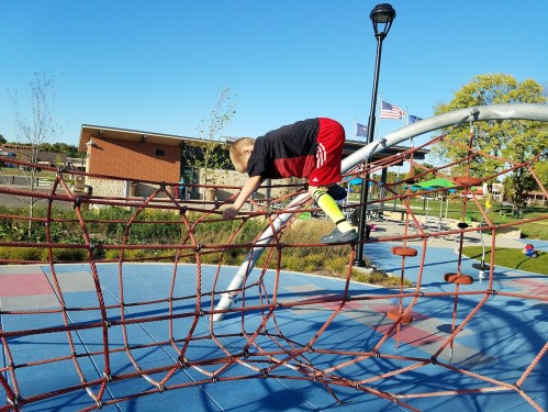 A child climbs on a playground climbing structure on a sunny day, with trees and a park in the background.