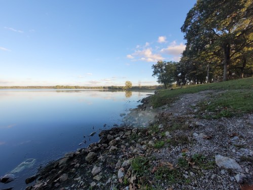 A serene lakeside view with calm water, rocky shore, and trees under a clear blue sky.