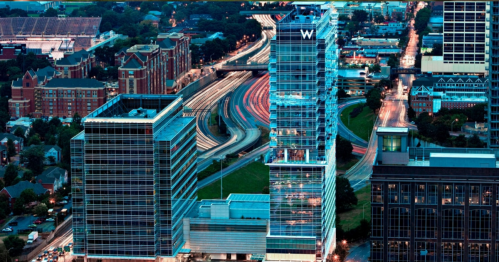 Aerial view of modern skyscrapers and a busy highway, illuminated at dusk with city lights in the background.