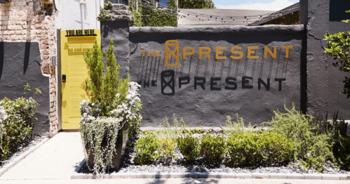 A vibrant wall with "THE PRESENT" and "PRESENT" painted in yellow, alongside a bright yellow door and greenery.