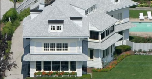 Aerial view of a large, white multi-story house with a gray roof and landscaped yard near a swimming pool.