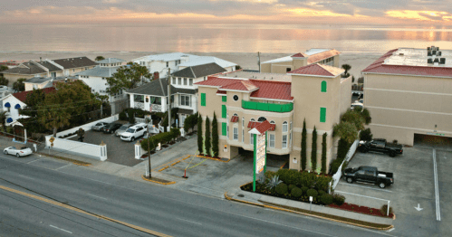 Aerial view of a beachfront hotel with a red roof, surrounded by palm trees and parked cars, near the ocean at sunset.