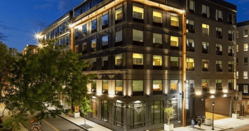 Modern building illuminated at night, featuring large windows and a sleek design, surrounded by trees and streetlights.