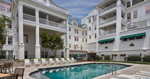 A serene pool area surrounded by white buildings and green trees, with lounge chairs arranged around the water.