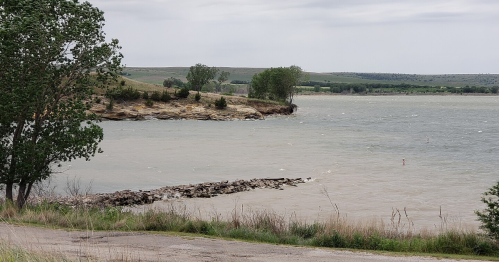 A serene lakeside view with rocky shorelines, green trees, and gentle waves under a cloudy sky.