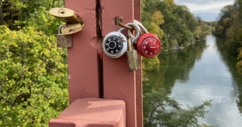 Two padlocks, one red and one white, hang on a bridge railing overlooking a calm river surrounded by trees.
