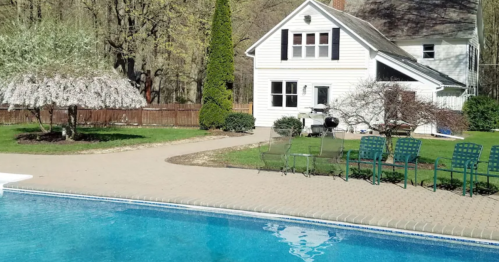A backyard with a swimming pool, a white house, and blooming trees, surrounded by green grass and patio chairs.