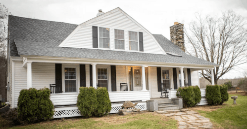 A charming white house with a stone chimney, rocking chairs on the porch, and a grassy yard.