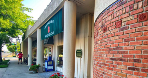 A brick building with a green awning, featuring a sign and flowers, alongside a sidewalk and a person walking.