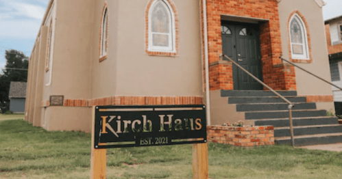 A building with arched windows and a sign reading "Kirch Haus EST. 2021" in front, surrounded by grass.