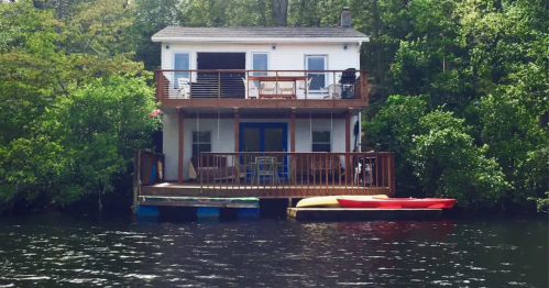 A cozy waterfront house with a deck, surrounded by greenery, and kayaks on the water.