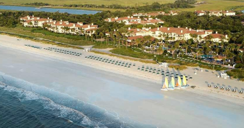 Aerial view of a beach resort with colorful sailboats, sun loungers, and lush greenery along the shoreline.