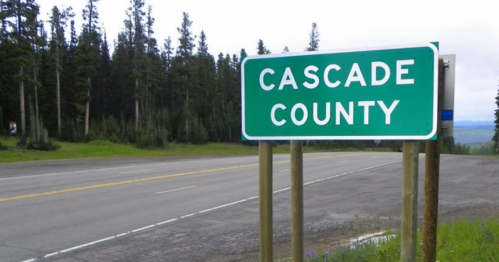 Green road sign for Cascade County beside a highway, surrounded by trees and open land.