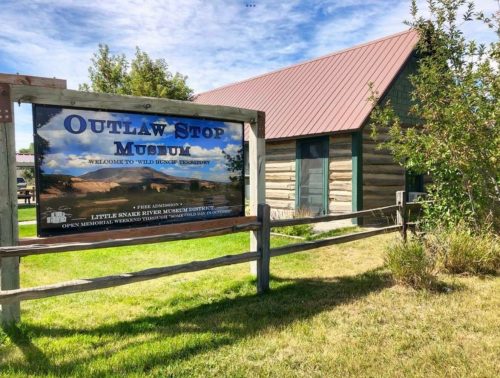 Sign for Outlaw Stop Museum with a rustic building in the background, surrounded by grass and trees.