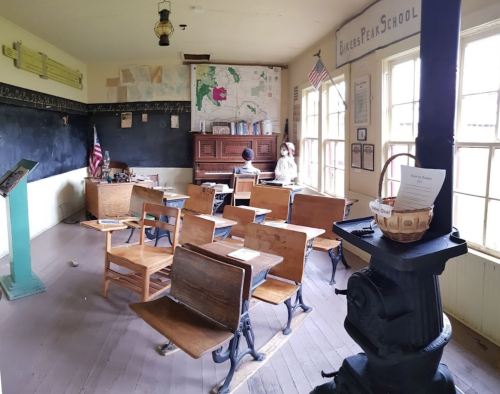 Historic classroom with wooden desks, a blackboard, a map, and a vintage stove, evoking a 19th-century school setting.