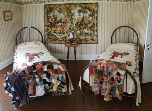 Two vintage beds with colorful quilts in a room featuring a tapestry and floral wallpaper. A small table is between the beds.