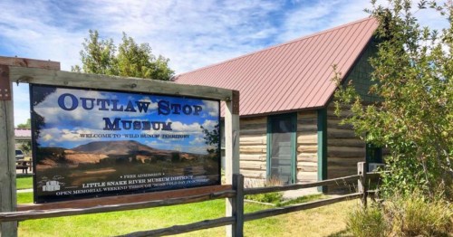Sign for Outlaw Stop Museum with a rustic building in the background, surrounded by grass and shrubs.
