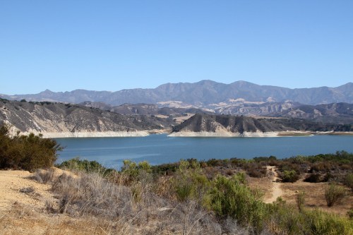 A serene lake surrounded by hills and mountains under a clear blue sky, with sparse vegetation in the foreground.