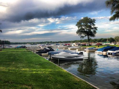 A serene lakeside view with boats docked, green grass, and a cloudy sky reflecting on the water.