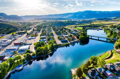 Aerial view of a scenic town by a river, surrounded by green hills and mountains under a bright blue sky.