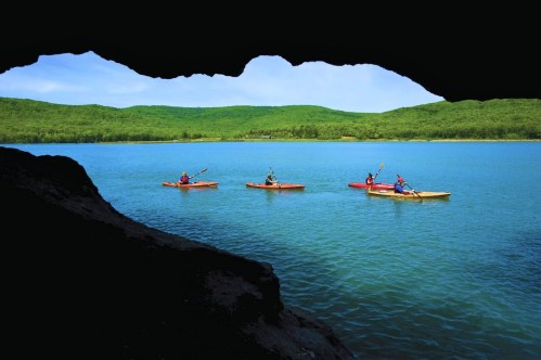 Four kayakers paddle on a serene lake, framed by a rocky cave entrance and lush green hills in the background.