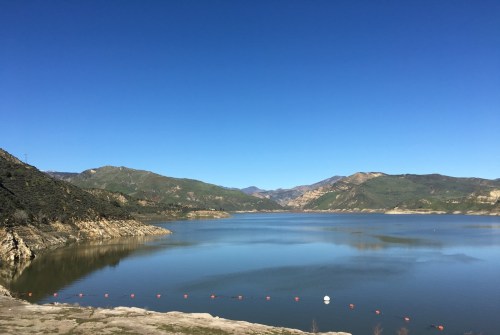 A serene lake surrounded by green hills under a clear blue sky, with distant mountains in the background.