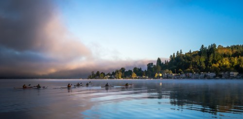 A serene lake scene at dawn with rowers gliding across the water and mist rising from the shore.