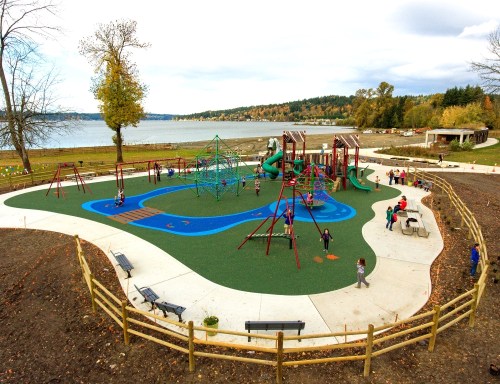 A playground by a lake, featuring slides, climbing structures, and children playing, surrounded by trees and benches.