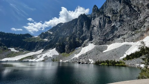 A serene mountain lake surrounded by rocky cliffs and patches of snow under a bright blue sky with fluffy clouds.