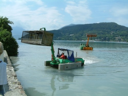 Two small boats on a river, one lifting a large container, surrounded by greenery and mountains in the background.