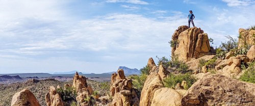 A hiker stands atop a rocky outcrop, overlooking a vast desert landscape under a blue sky with scattered clouds.